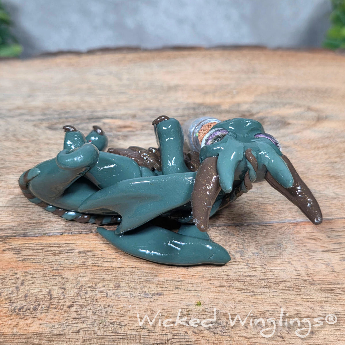 A clay dragon with a potion bottle on a wood surface. Text reads Wicked Winglings.