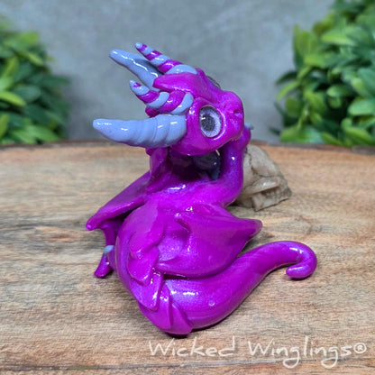  A clay dragon holding a skull sitting against a blurred background with greenery. Text says Wicked Winglings.