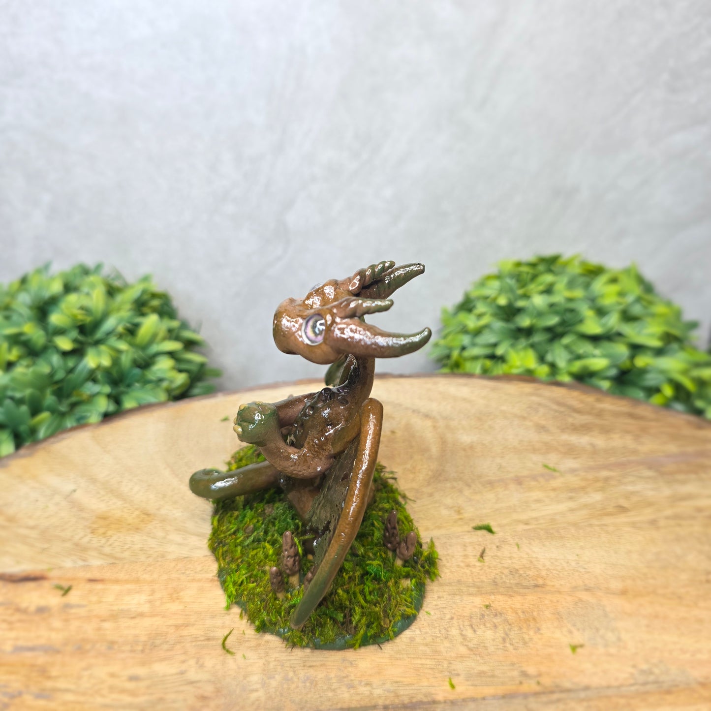 Small dragon figurine on a piece of wood with greenery in the background