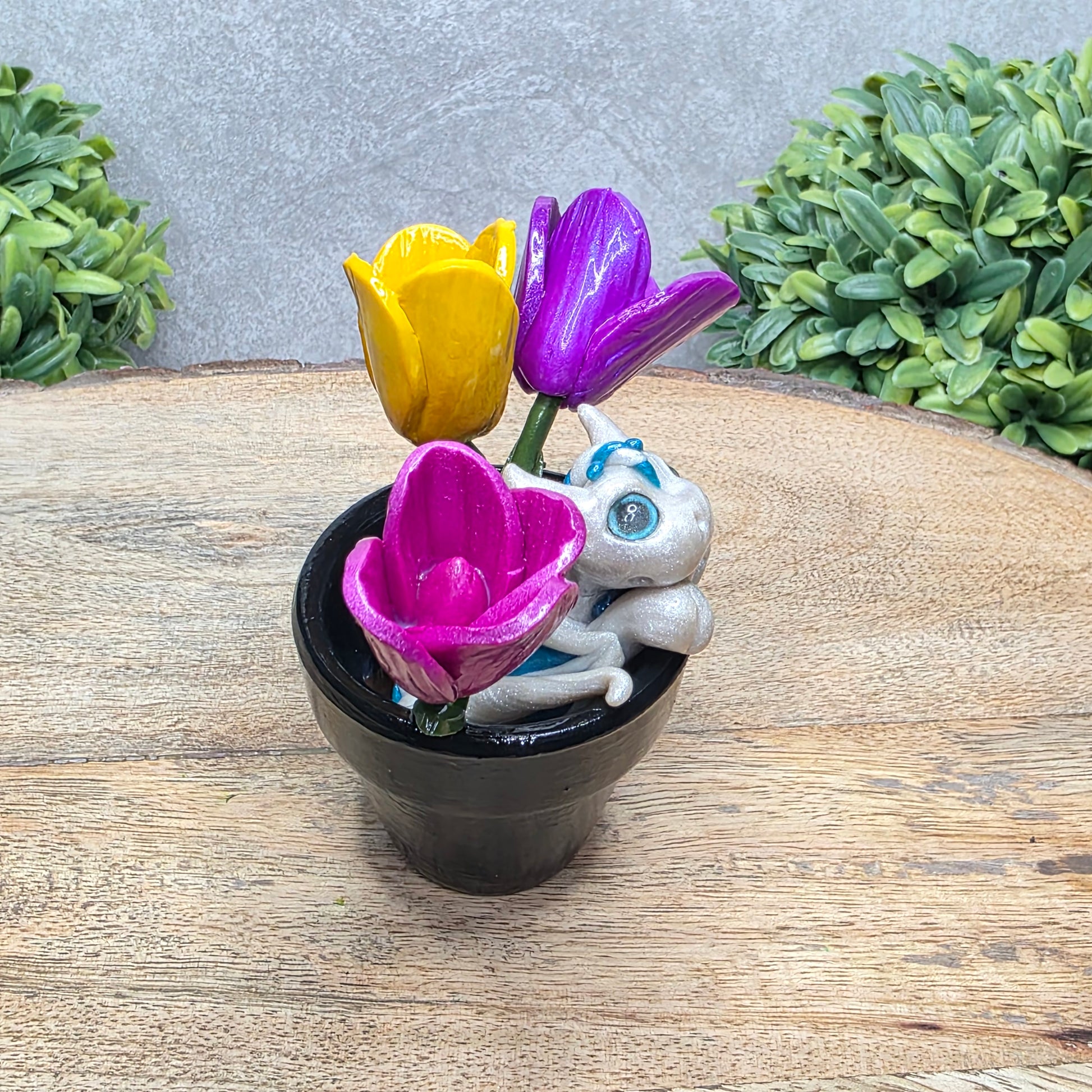 Side view of a clay baby dragon in a pot with tulips against a wood surface with greenery.