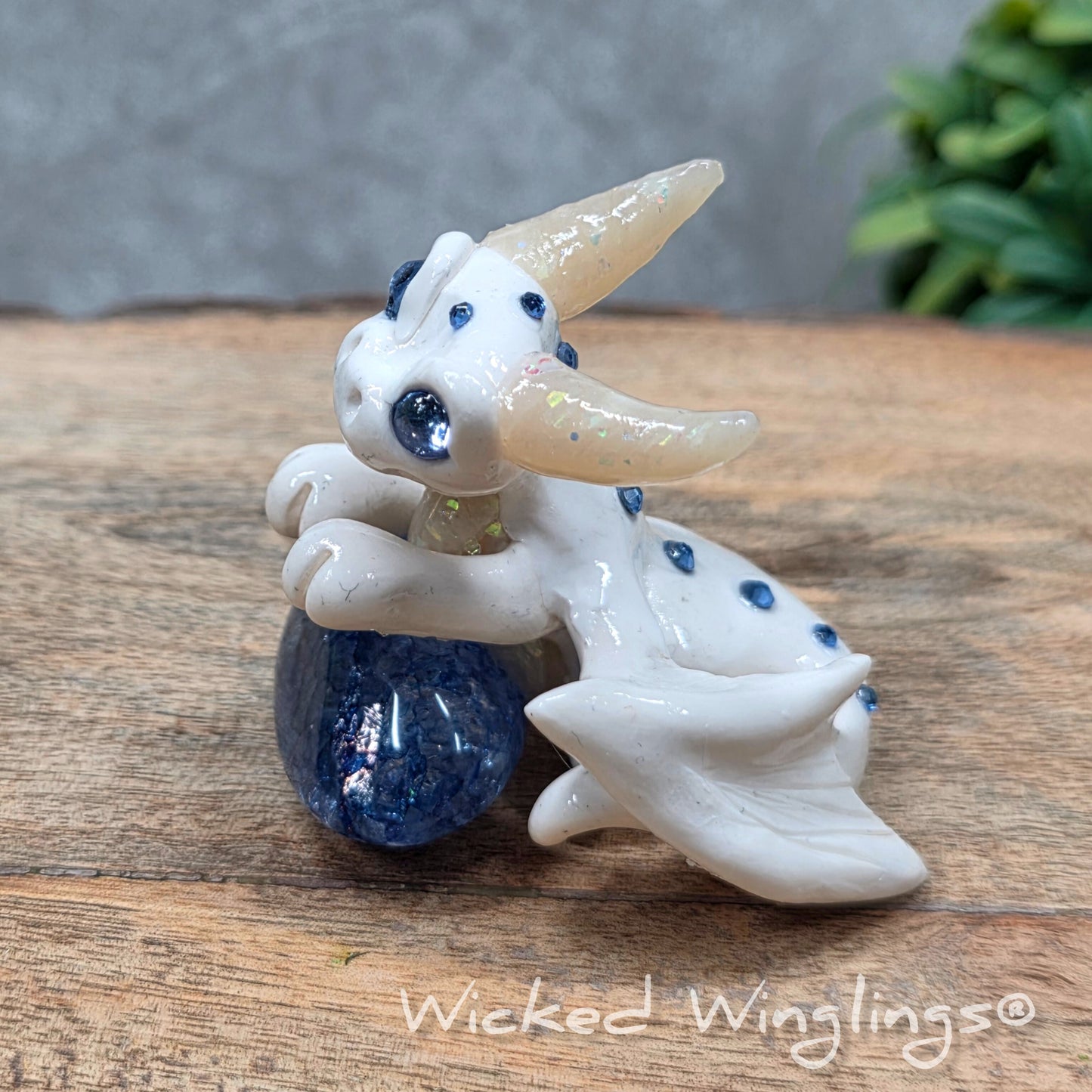 A baby clay dragon sculpture with a crackled quartz against a wood background with greenery and text Wicked Winglings