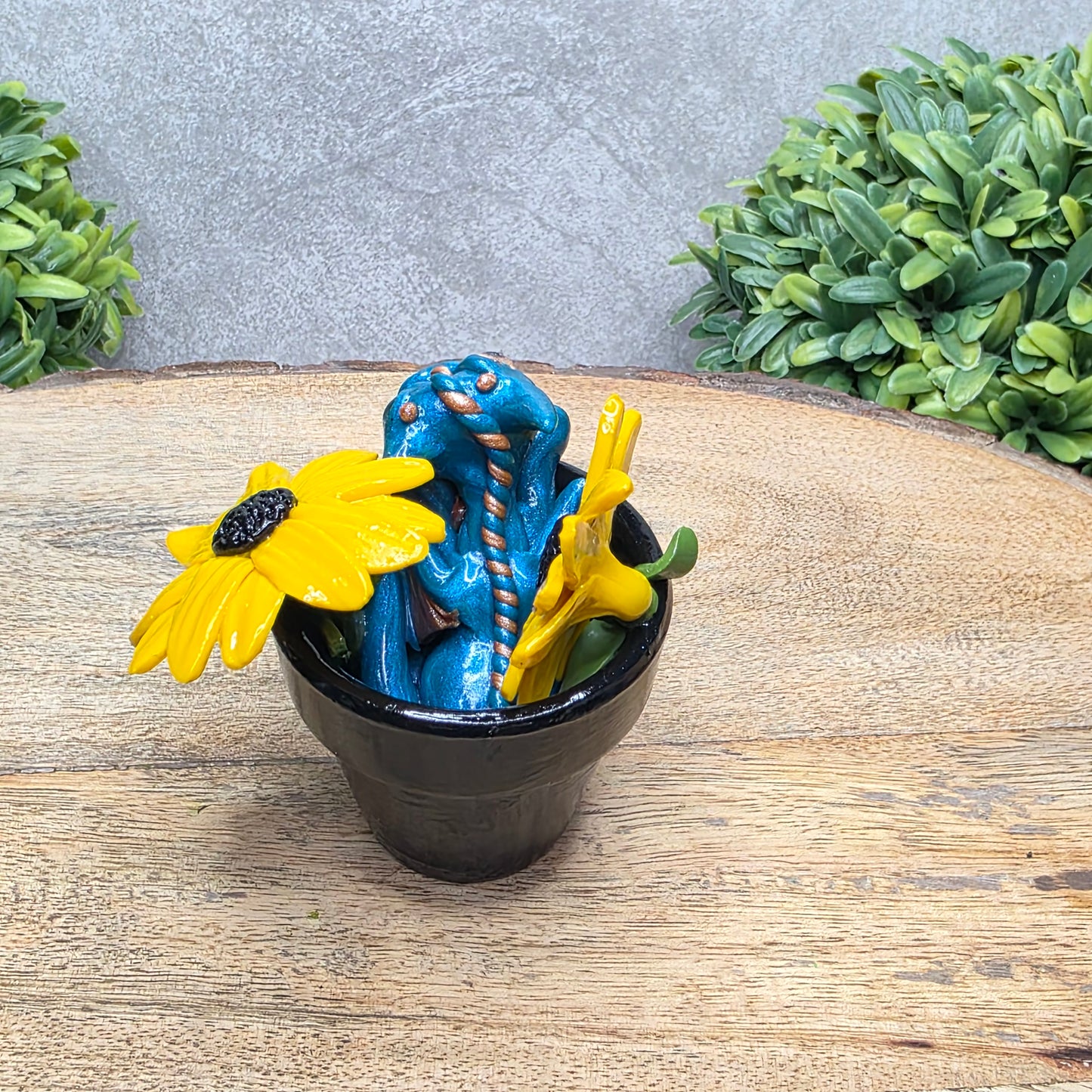 Back view of a clay baby dragon in a pot with black eye susans against a wood surface with greenery.