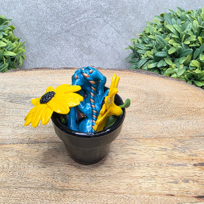 Back view of a clay baby dragon in a pot with black eye susans against a wood surface with greenery.