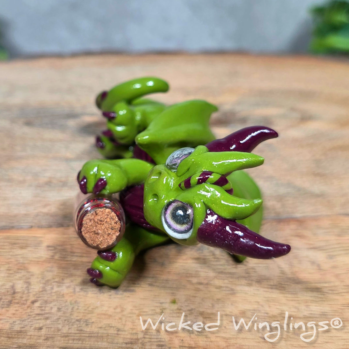 A clay dragon with a potion bottle on a wood surface. Text reads Wicked Winglings.