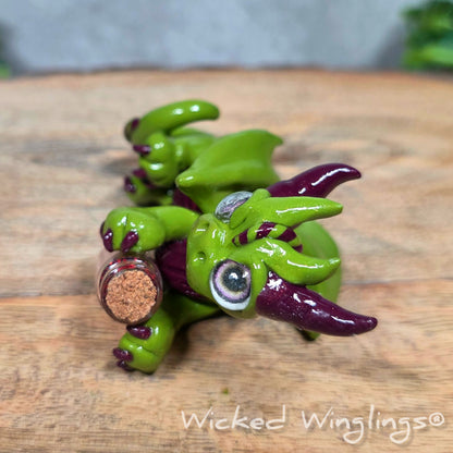 A clay dragon with a potion bottle on a wood surface. Text reads Wicked Winglings.