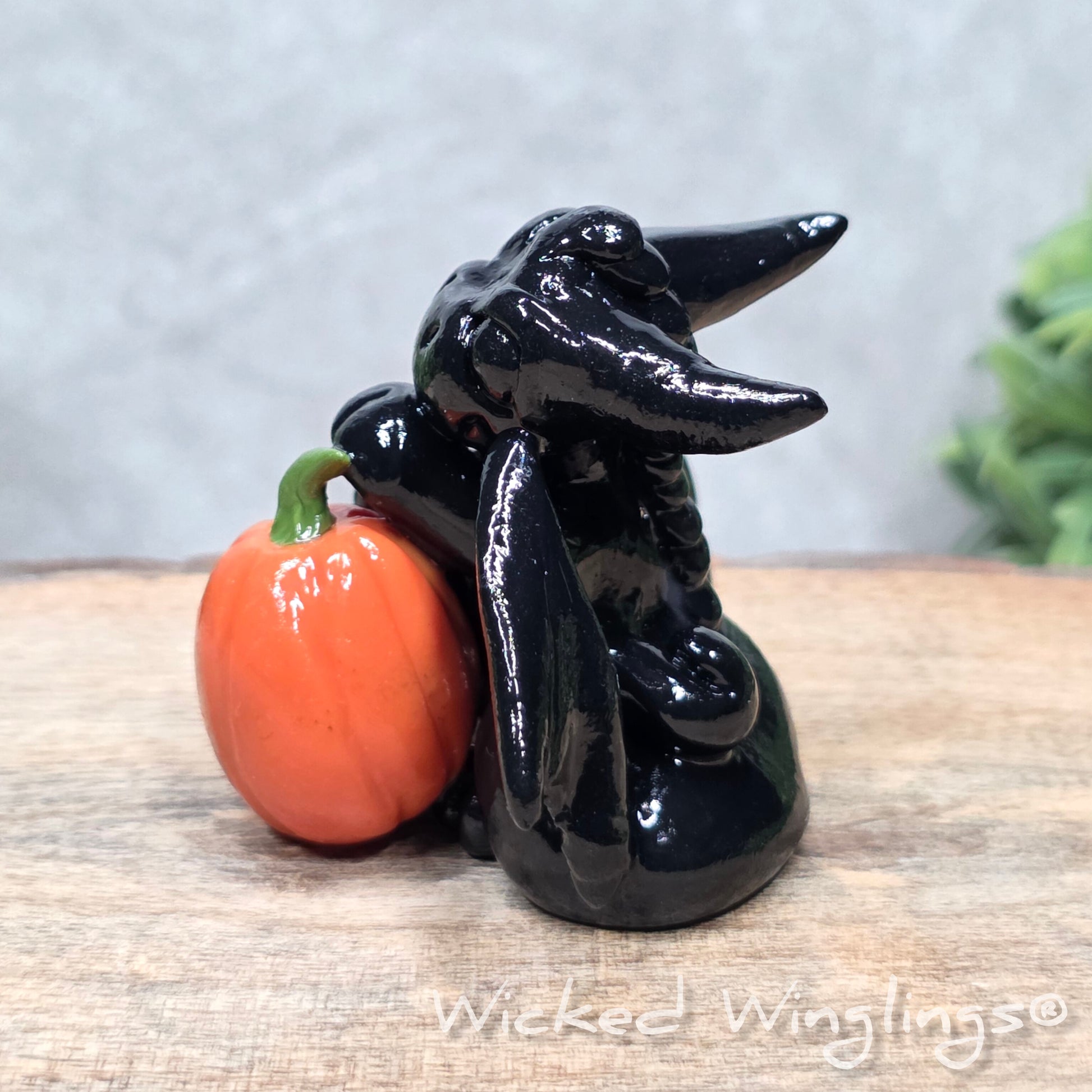 A baby clay dragon sculpture with pumpkins against a wood background with greenery and text Wicked Winglings