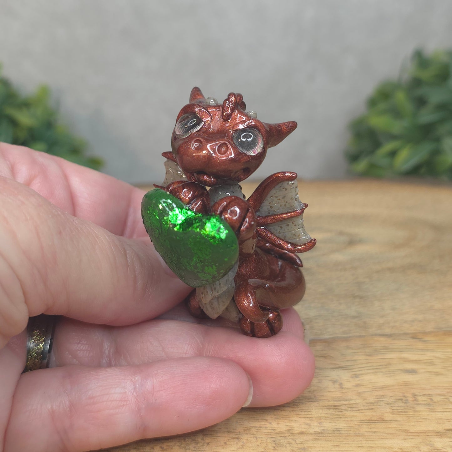 A hand holding a baby polymer clay dragon with a green heart against a blurred background
