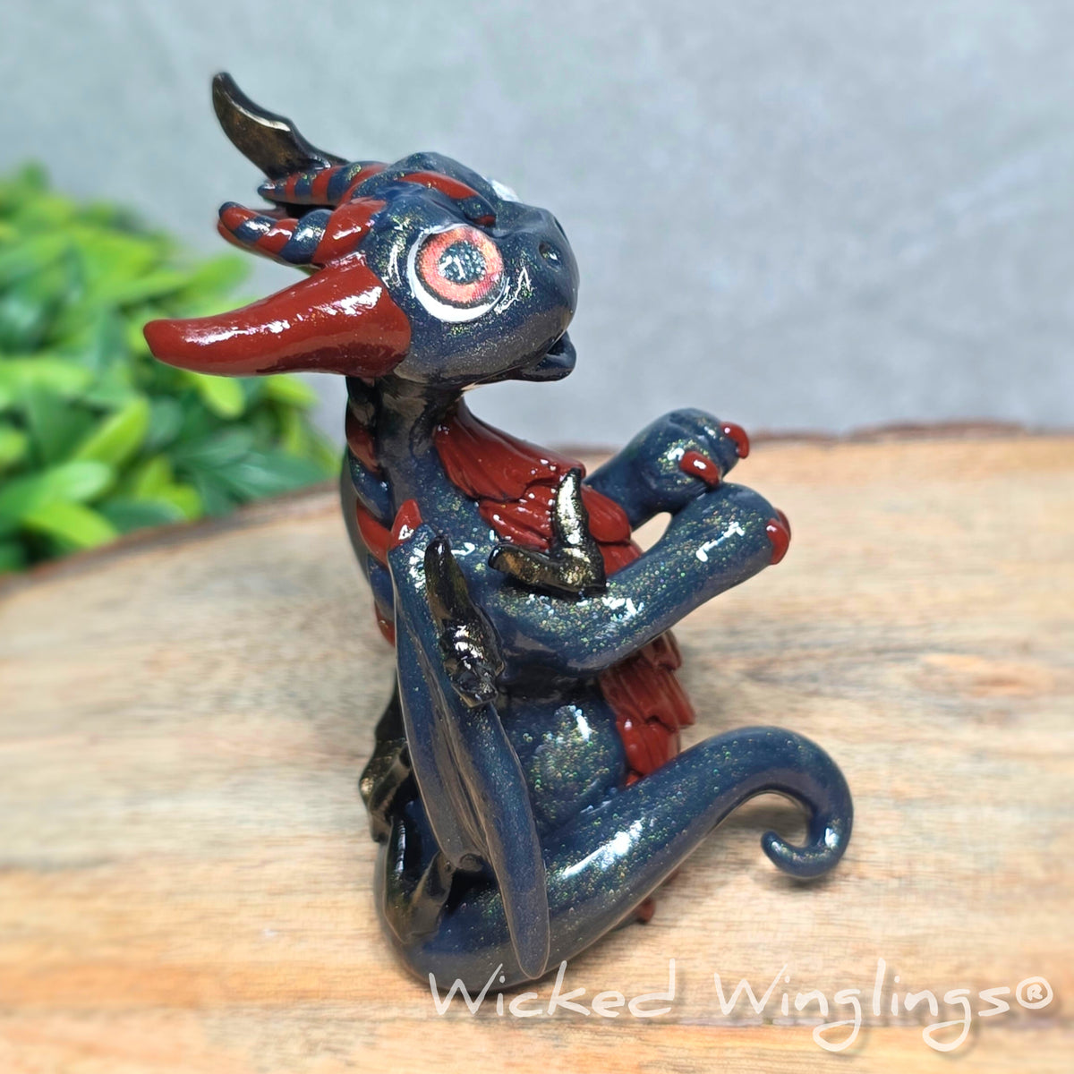 A side view of a clay dragon with bats sitting on a wood surface with greenery. Text reads Wicked Winglings.