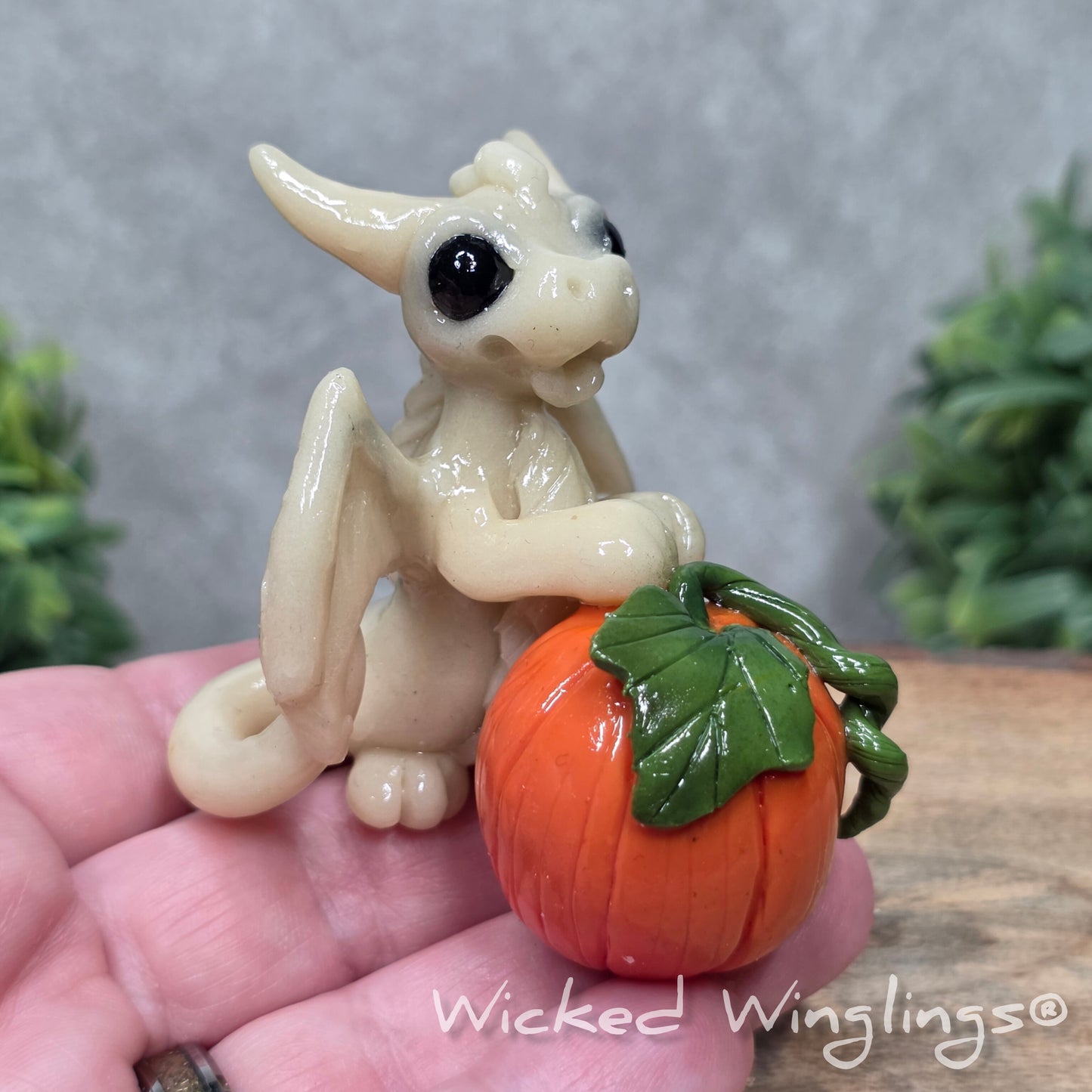A hand holding a baby clay dragon  sculpturewith a pumpkin against a wood background with greenery and text Wicked Winglings