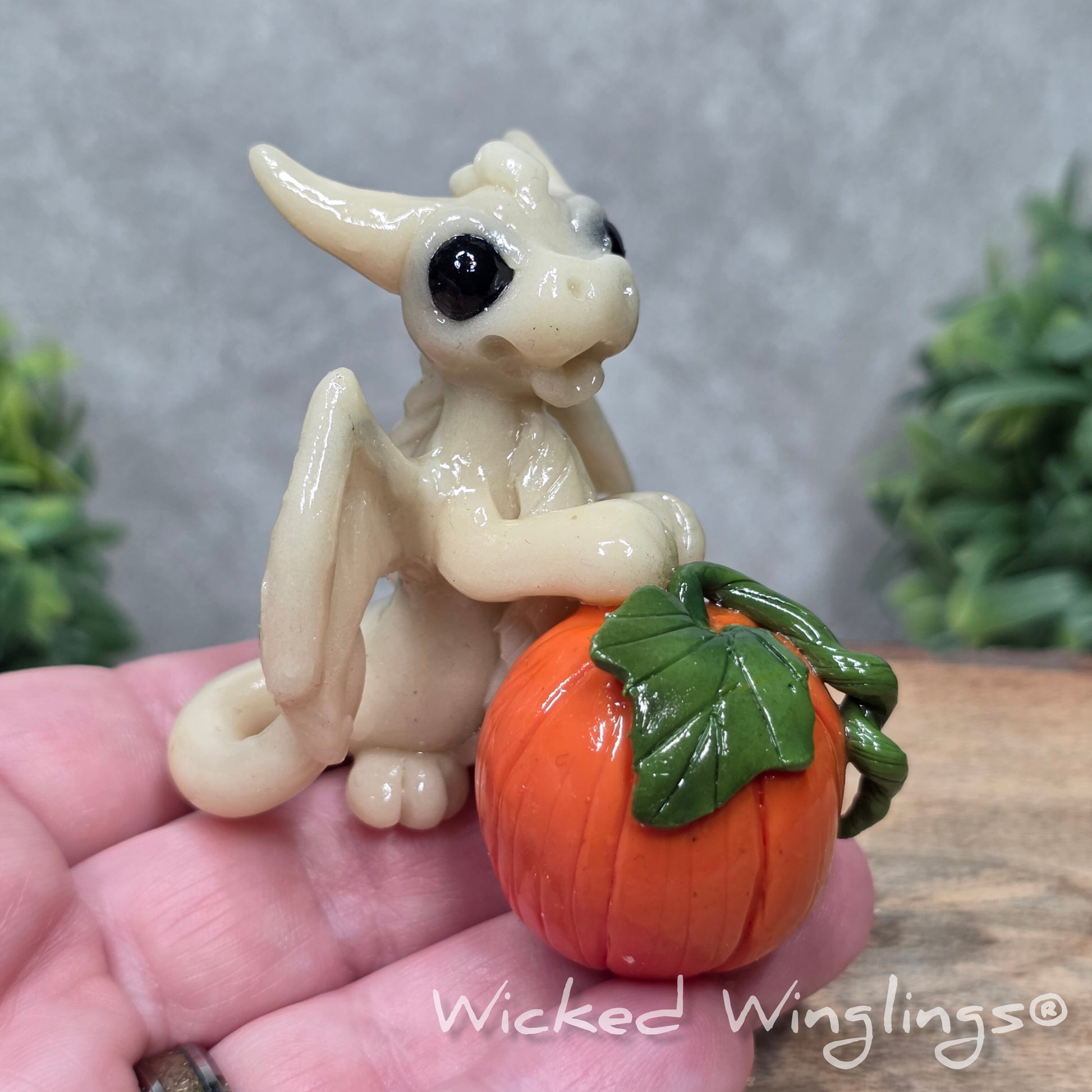 A hand holding a baby clay dragon  sculpturewith a pumpkin against a wood background with greenery and text Wicked Winglings