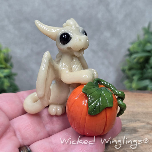 A hand holding a baby clay dragon  sculpturewith a pumpkin against a wood background with greenery and text Wicked Winglings