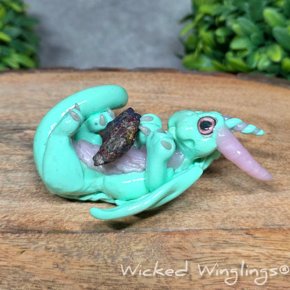 A mint clay dragon holding a rainbow colored stone against a blurred background with greenery. Text says Wicked Winglinng.