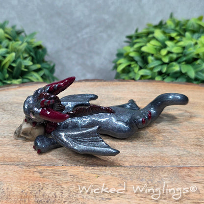 A clay dragon with a skull against a wood surface with greenery. Includes text Wicked Winglings