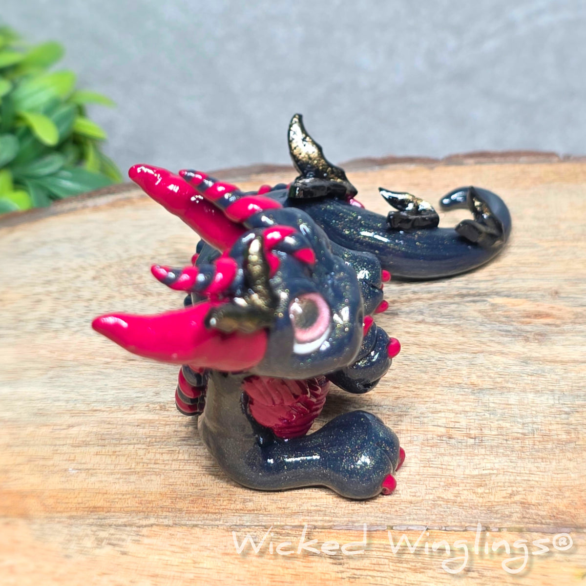 Side view of a clay dragon with bats sitting on a wood surface with greenery. Text reads Wicked Winglings.