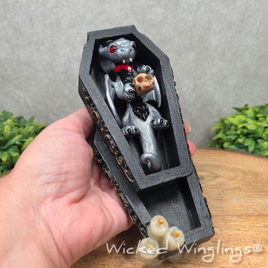 A hand holding a clay dragon in a black wood coffin with candles and a skull