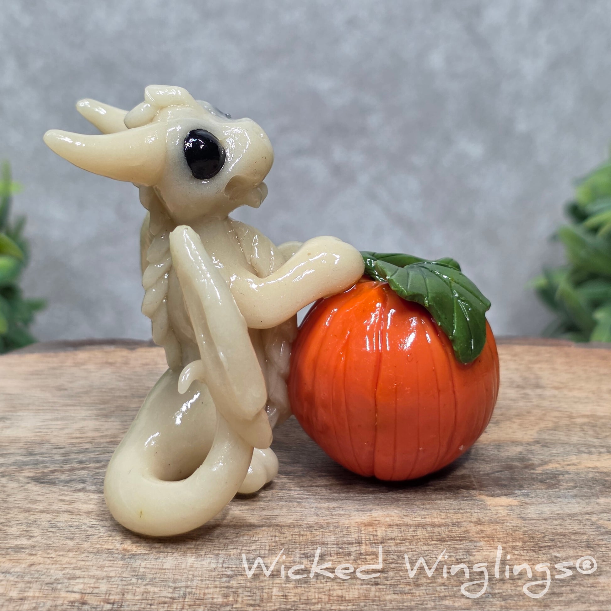 A baby clay dragon sculpture with a pumpkin against a wood background with greenery and text Wicked Winglings