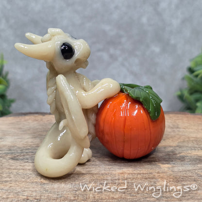 A baby clay dragon sculpture with a pumpkin against a wood background with greenery and text Wicked Winglings
