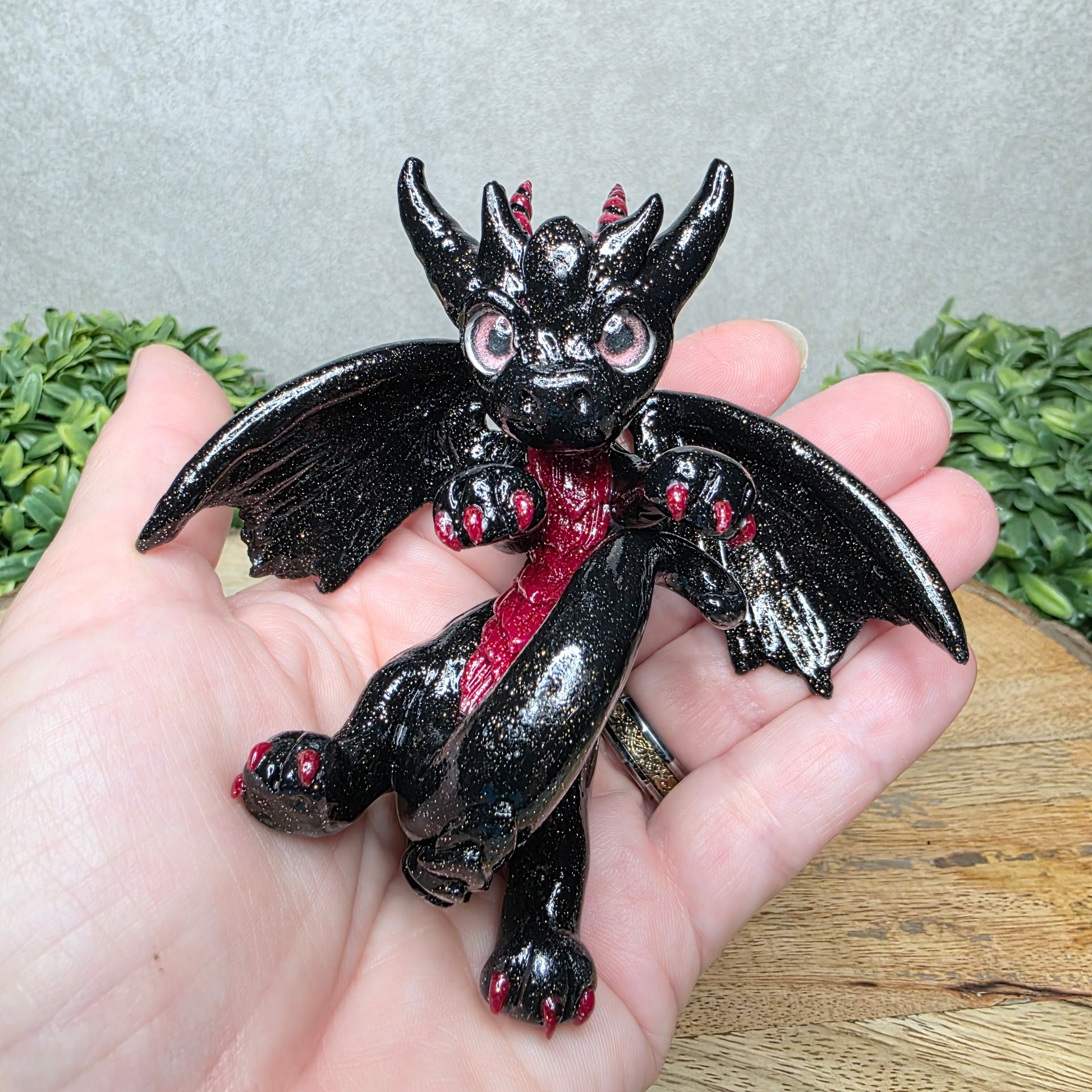 A hand holding a clay dragon against a wood background with greenery
