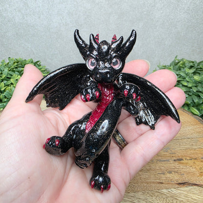 A hand holding a clay dragon against a wood background with greenery