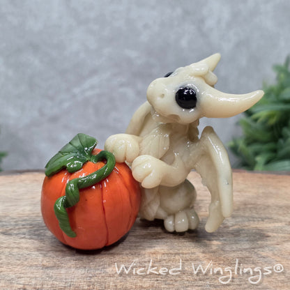 A baby clay dragon sculpture with a pumpkin against a wood background with greenery and text Wicked Winglings