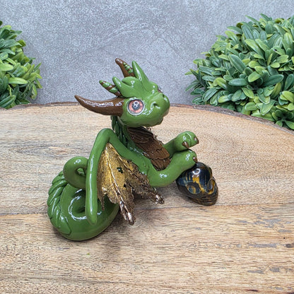 Side view of a clay dragon with a tigers eye crystal skull against a wood surface with greenery.