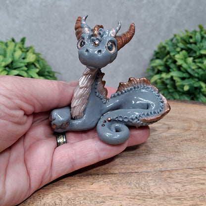 A hand holding a clay dragon against a wood background with greenery