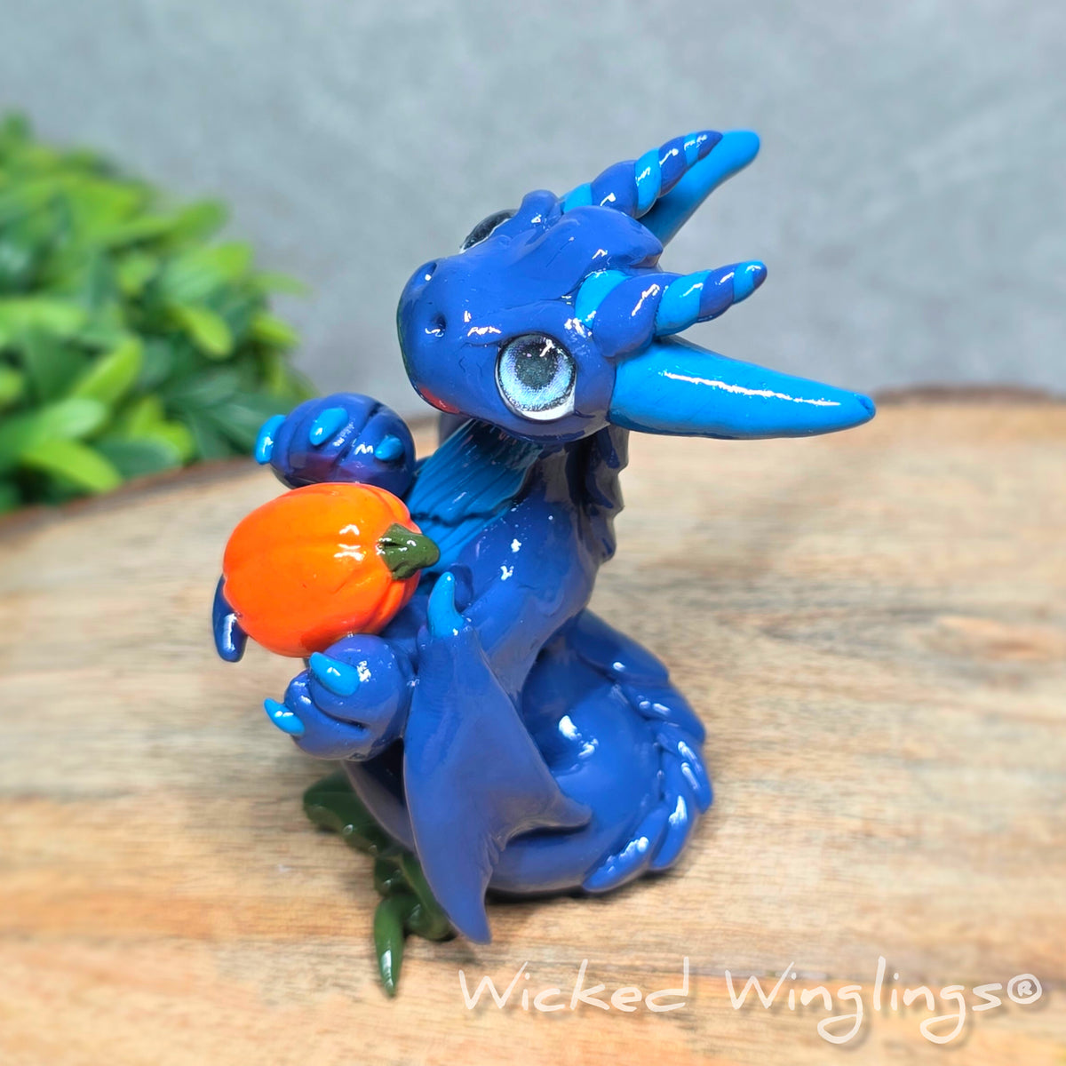 Side view of a blue clay dragon with pumpkins against a wood surface with greenery. Text reads Wicked Winglings. 