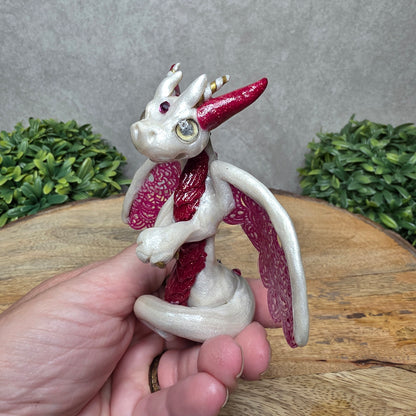 A hand holding a clay dragon against a wood background with greenery