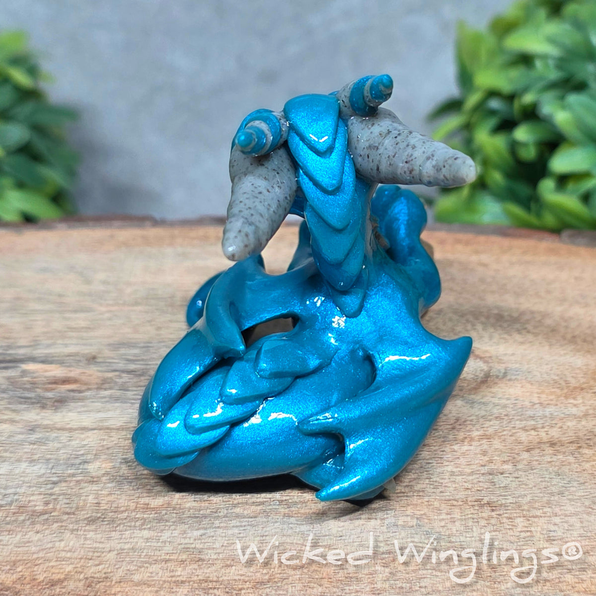  A clay dragon holding a skull sitting against a blurred background with greenery. Text says Wicked Winglings.