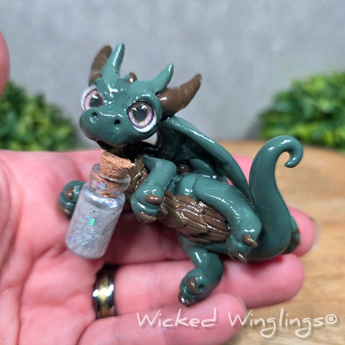 A hand holding a clay dragon with a potion bottle against a wood surface. Text reads Wicked Winglings.