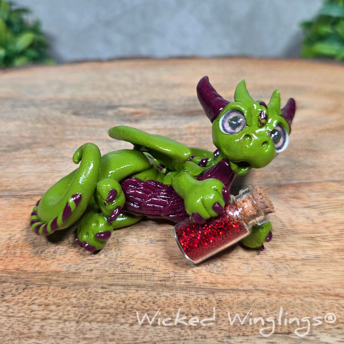 A clay dragon with a potion bottle on a wood surface. Text reads Wicked Winglings.