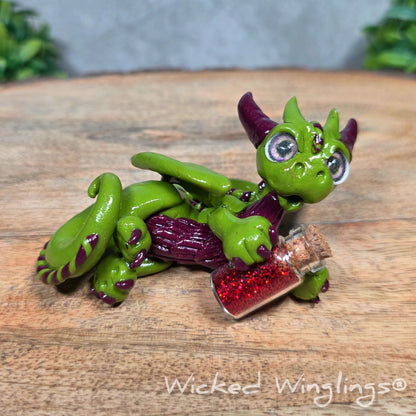 A clay dragon with a potion bottle on a wood surface. Text reads Wicked Winglings.