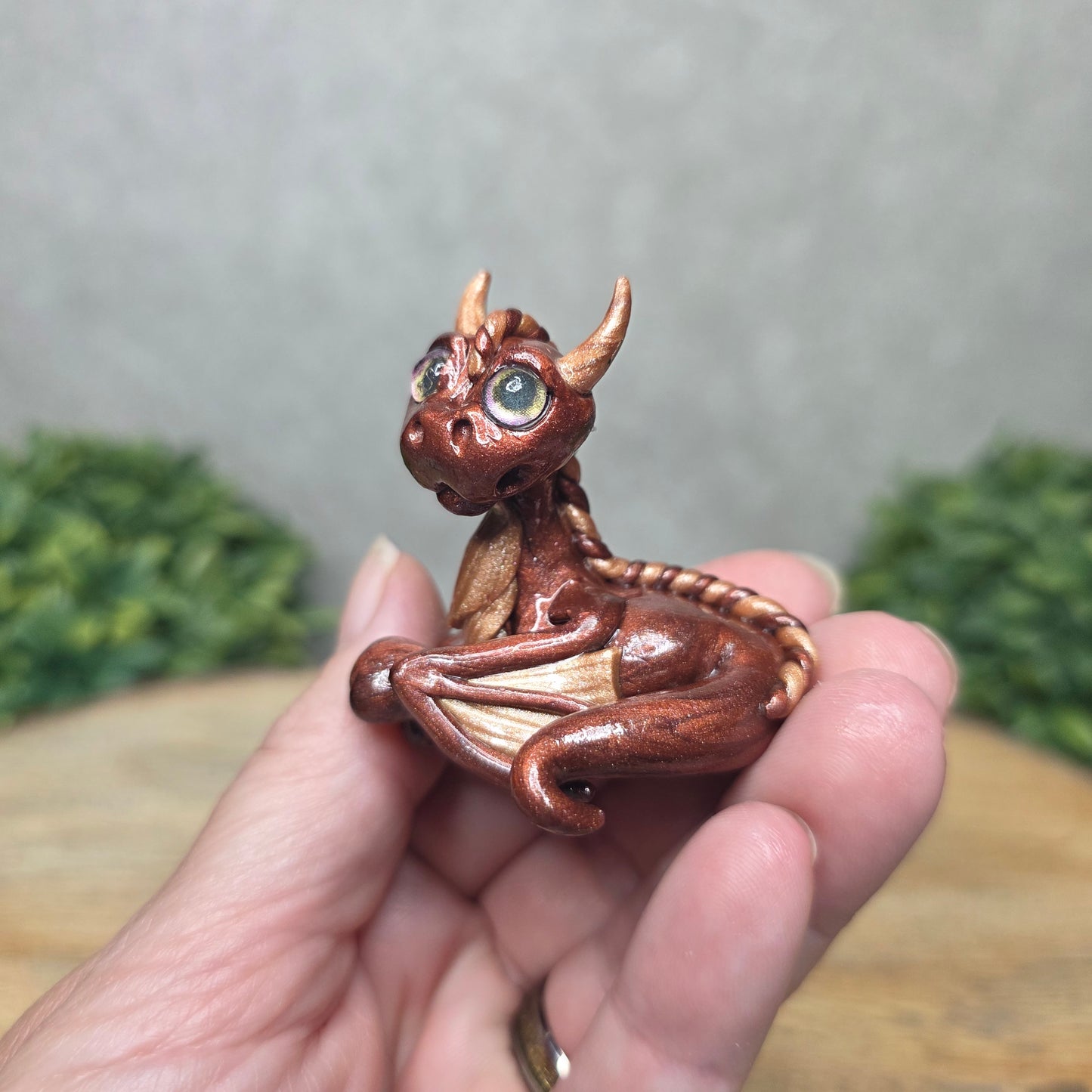 A hand holding a baby clay dragon against a wood background with greenery