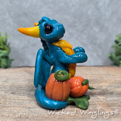 A baby clay dragon sculpture with pumpkins against a wood background with greenery and text Wicked Winglings