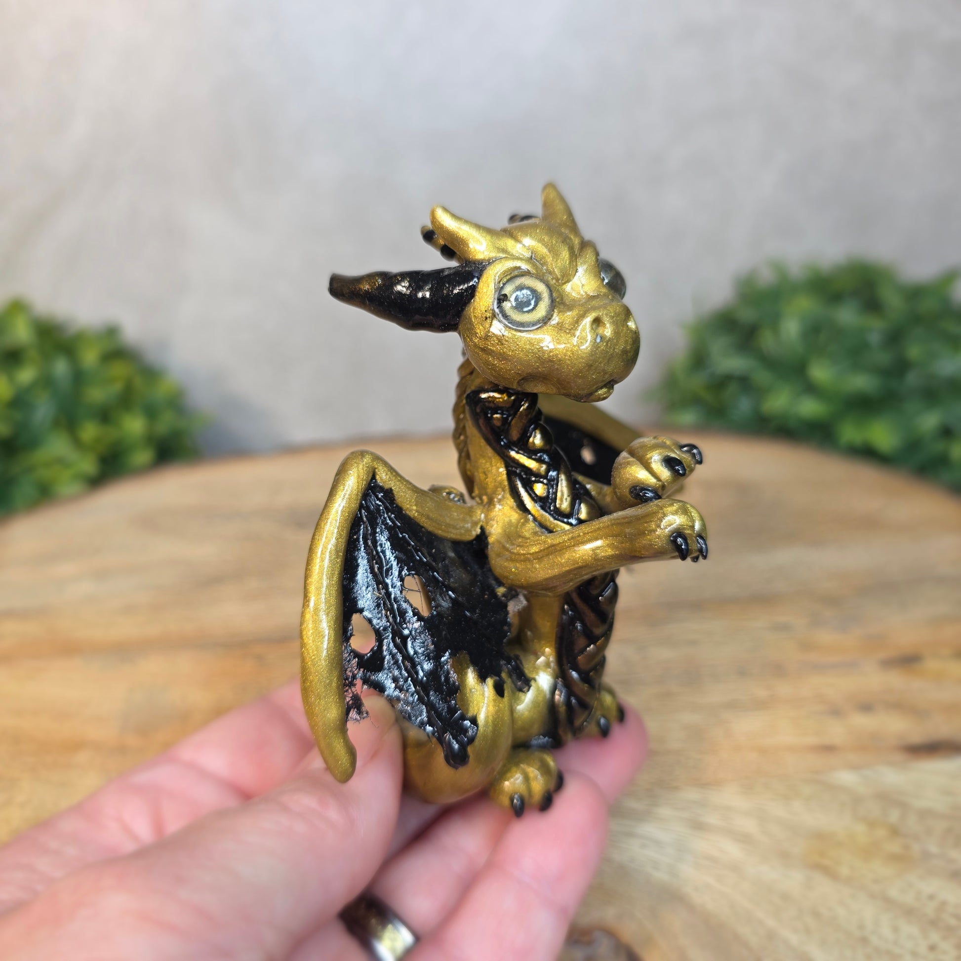 A hand holding a clay dragon figurine on a wooden surface with a blurred green plant in the background