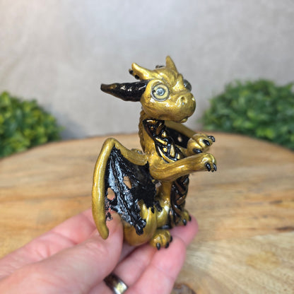 A hand holding a clay dragon figurine on a wooden surface with a blurred green plant in the background