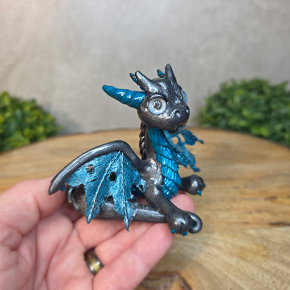 A hand holding a graphite and blue clay dragon against a blurred wood surface