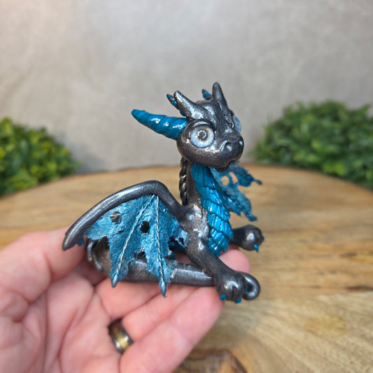A hand holding a graphite and blue clay dragon against a blurred wood surface