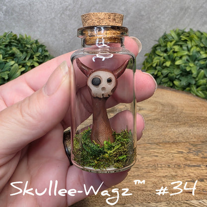 Hand holding a skull faced creature in a jar against a wood background with greenery