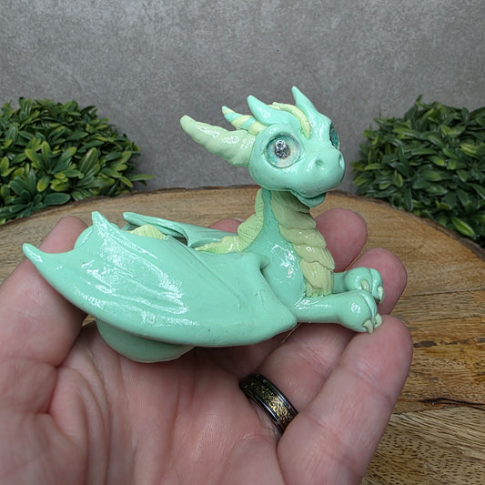 A hand holding a clay dragon against a wood background with greenery