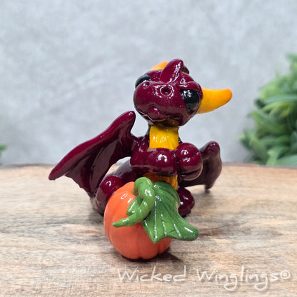 A baby clay dragon sculpture with a pumpkin against a wood background with greenery and text Wicked Winglings