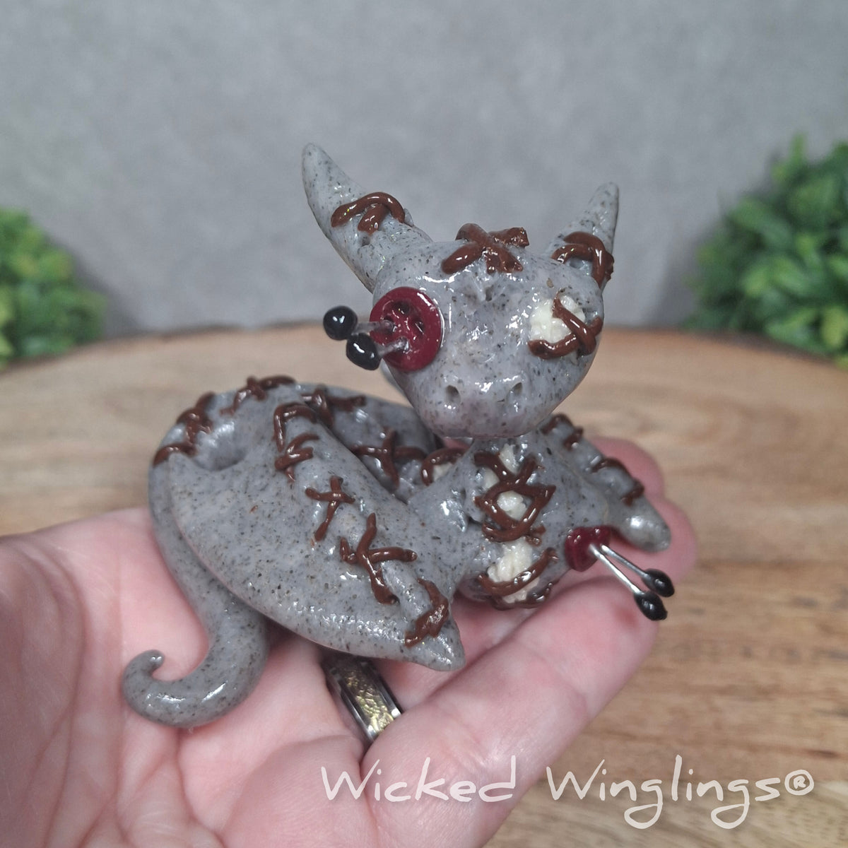 A hand holding a clay voodoo dragon against a wood surface with greenery. Text says Wicked Winglings