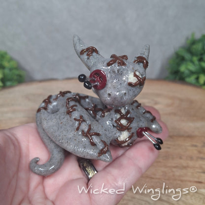 A hand holding a clay voodoo dragon against a wood surface with greenery. Text says Wicked Winglings