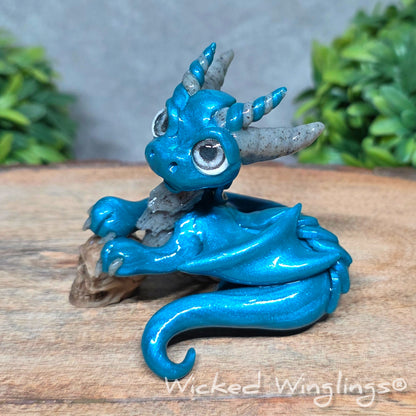  A clay dragon holding a skull sitting against a blurred background with greenery. Text says Wicked Winglings.
