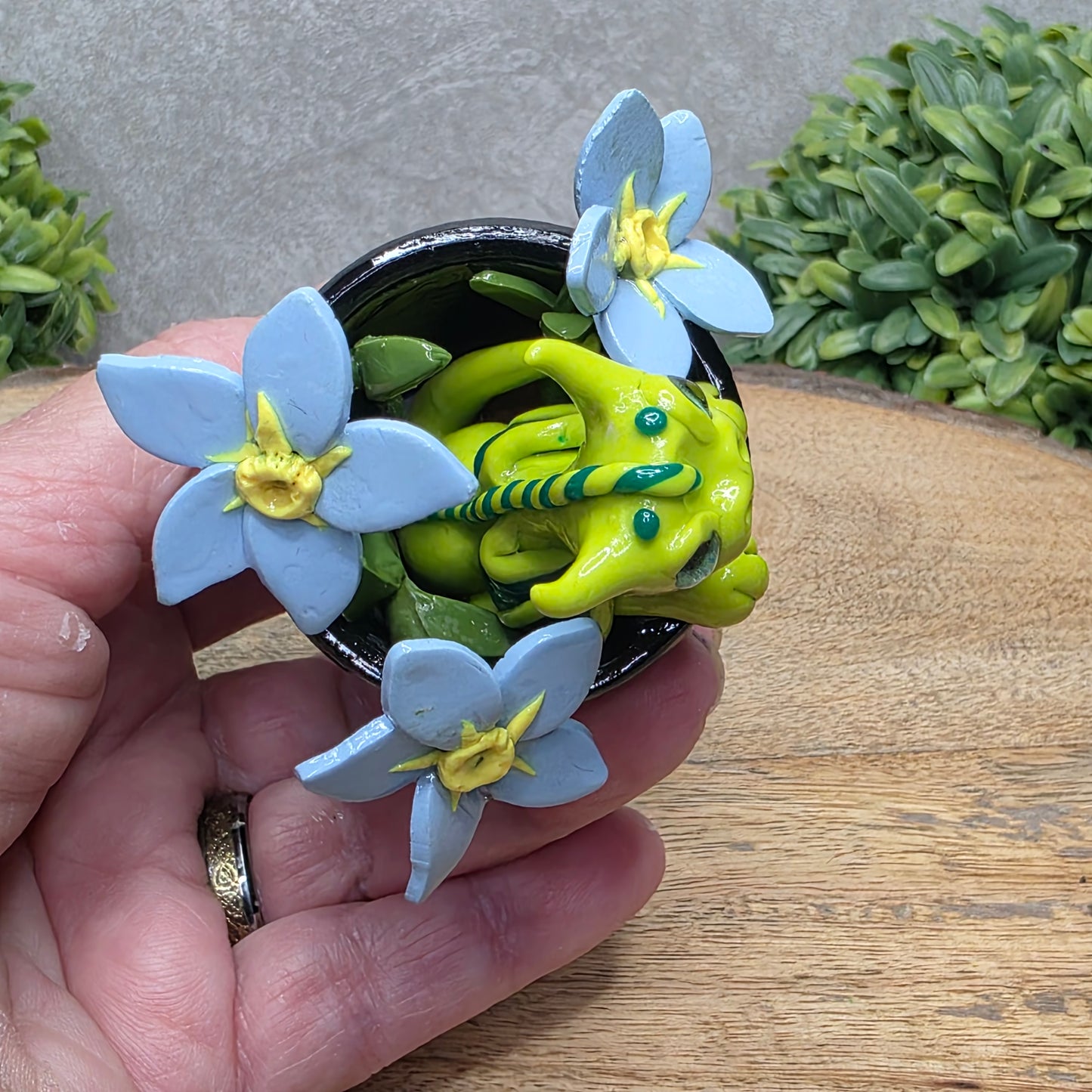 Top view of a hand holding a clay baby dragon in a pot with forget me nots against a wood surface with greenery.