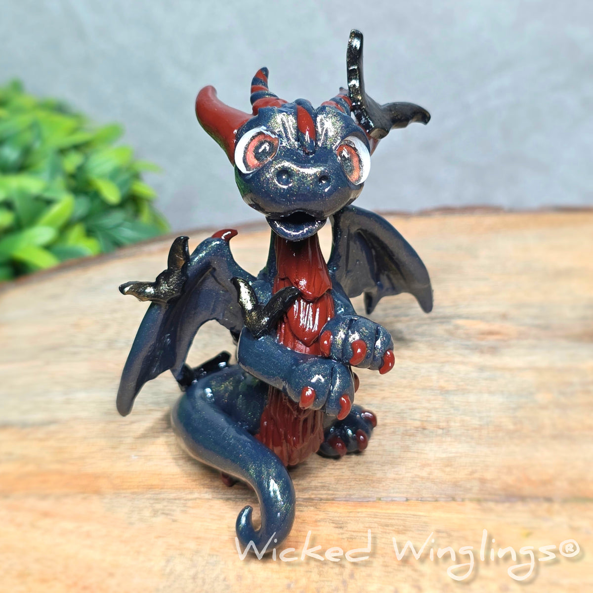 A clay dragon with bats sitting on a wood surface with greenery. Text reads Wicked Winglings.