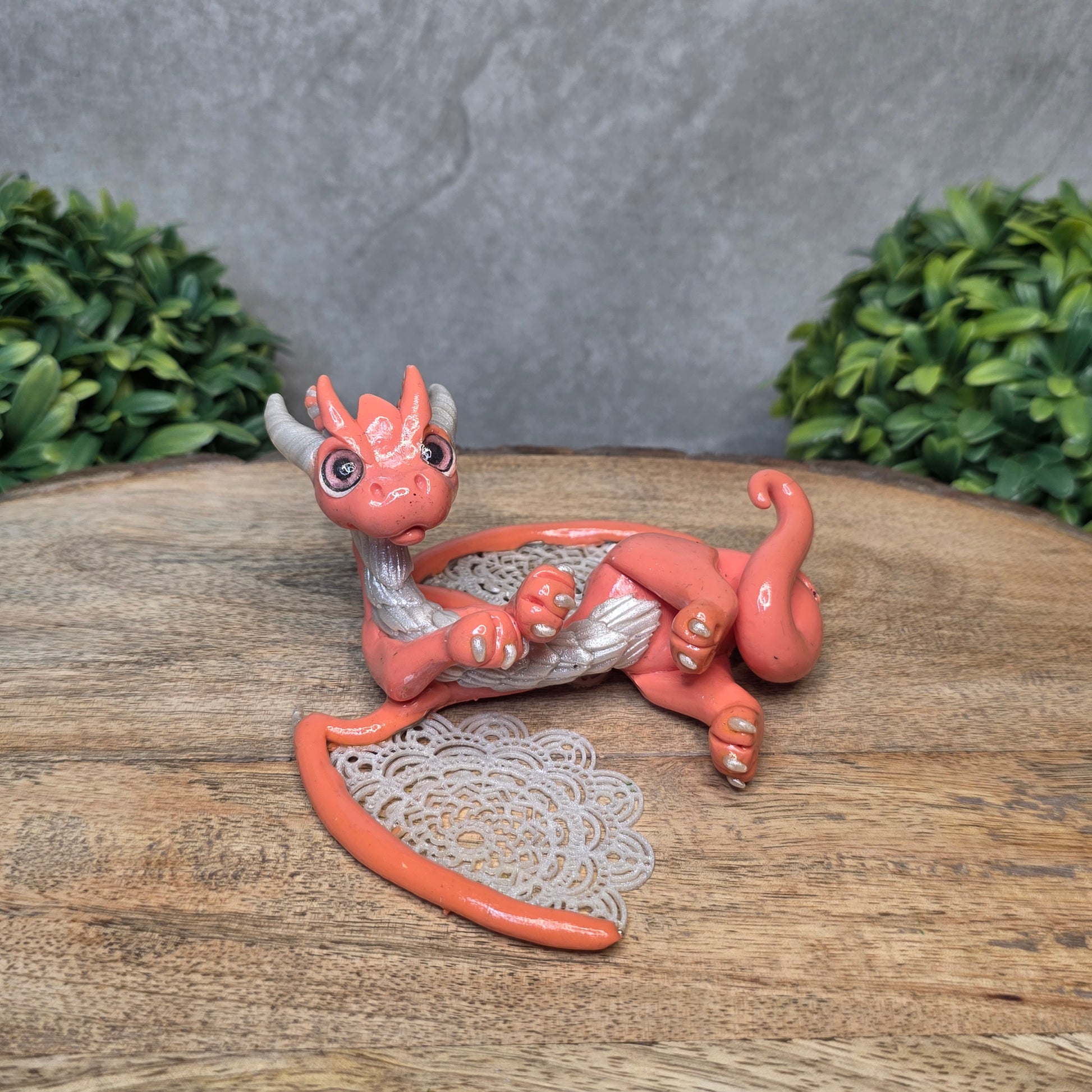 A clay dragon sculpture with lacy wings on a wood surface with greenery.