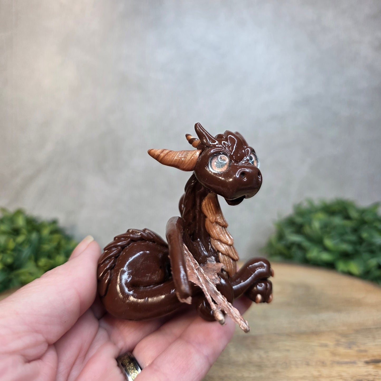 A hand holding a brown and copper clay dragon against a blurred wood surface