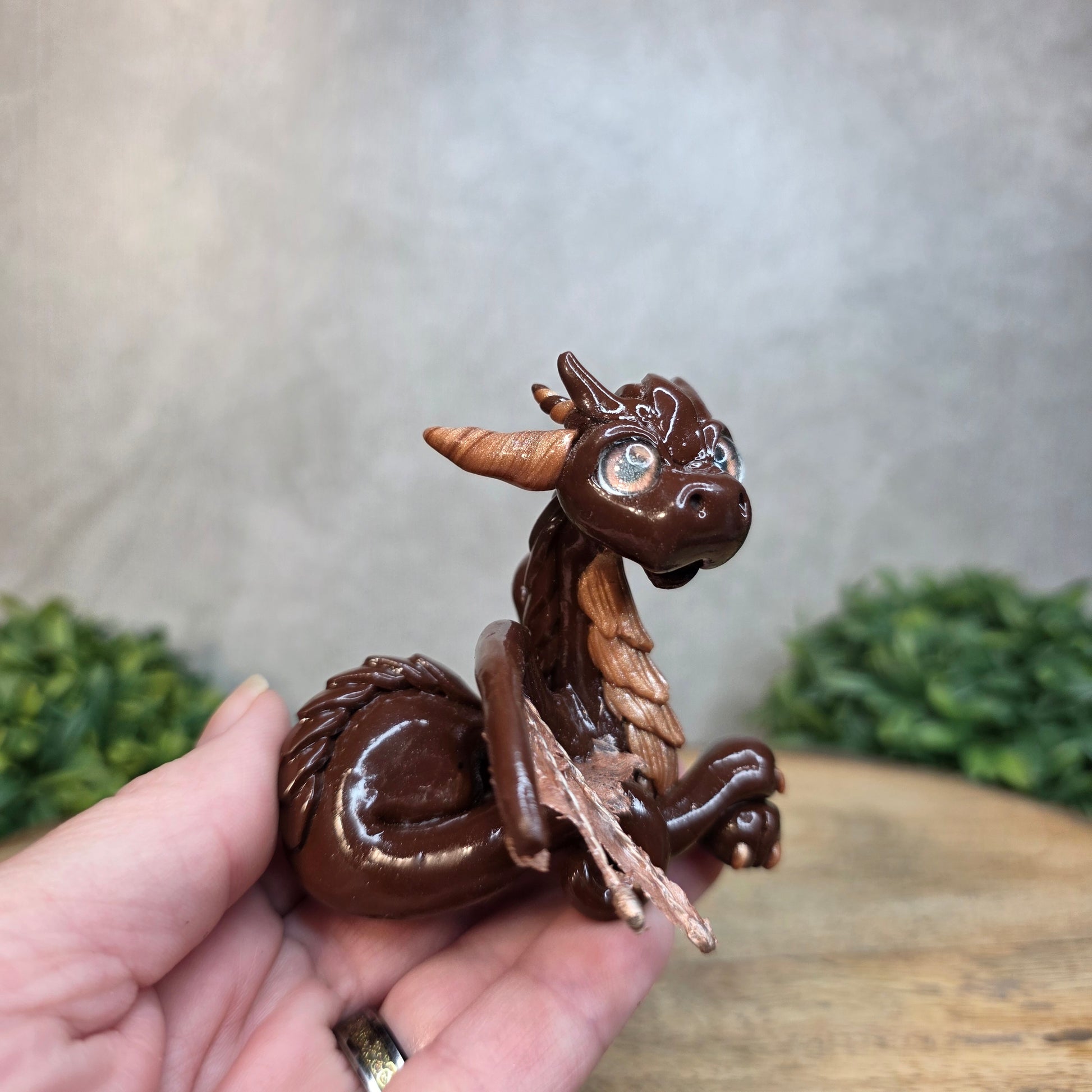 A hand holding a brown and copper clay dragon against a blurred wood surface