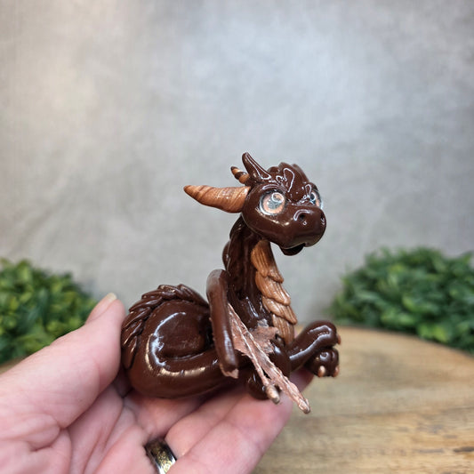A hand holding a brown and copper clay dragon against a blurred wood surface